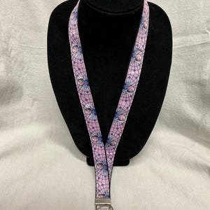 May include: A pink lanyard with a repeating pattern of dream catchers and spiderwebs. The lanyard has a silver-colored metal clasp and key ring. The design features blue and white accents on a light pink background. The lanyard is displayed on a black mannequin.