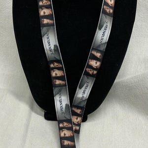 May include: A lanyard featuring a repeating pattern of faces and the text "SUPERNATURAL" in white on a gray background. The lanyard has a silver-colored metal clasp and a hook.