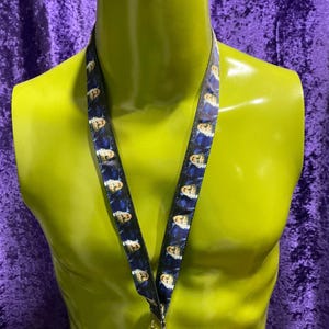 May include: A lanyard with a repeating pattern of a face on a dark blue background. The lanyard is displayed on a yellow mannequin. The lanyard has a gold-colored clasp.