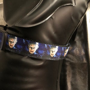 May include: A black dog collar with a blue and black patterned ribbon. The ribbon features a repeating image of a person's face. The collar is displayed on a black mannequin.