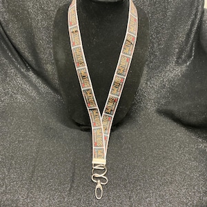 May include: A white lanyard with the word "SUBLIME" printed repeatedly in a black, red, and yellow design. The lanyard has a silver-colored metal clasp and is displayed on a black mannequin head.