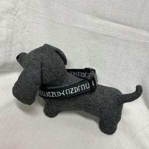 May include: A stuffed dachshund dog in gray herringbone fabric with a black collar. The collar has white text that reads "OZDUX KOZDROU". The dog has floppy ears and a small tail.