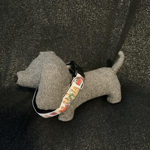 May include: A gray dachshund dog-shaped plush toy with a herringbone pattern. The dog has a black collar with a colorful pattern. The toy is on a black, glittery surface.
