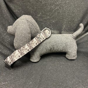 May include: A gray stuffed dachshund dog with a matching patterned collar. The dog is made of a herringbone fabric. The collar has a silver metal buckle and a black and white pattern. The background is a dark, glittery fabric.