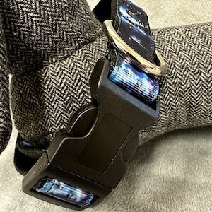 May include: A black dog collar with a cityscape design in blue, white, and purple. The collar has a black plastic buckle and a silver metal D-ring for leash attachment. The collar is shown against a gray background.