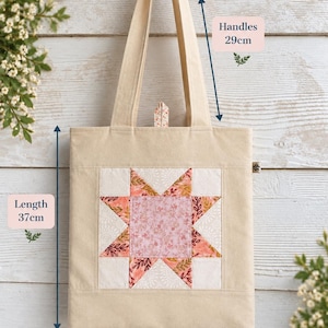 May include: A beige tote bag featuring a central star-shaped quilt block pattern in shades of pink, orange, and white. The bag has a length of 37 cm, a width of 31 cm, and handles measuring 29 cm. A small fabric tab is visible at the top centre.
