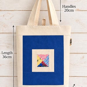 May include: A cream-coloured tote bag with a central blue rectangular panel and a small, multi-coloured floral square patch. The bag features two handles, a length of 36cm, a width of 32cm, a depth of 8cm, and a handle drop of 20cm.