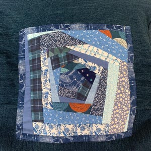 May include: A square fabric patch featuring a spiral patchwork design in various shades of blue, white, and orange. The quilt block includes patterns of flowers, plaid, and a bird illustration, all stitched onto a dark blue textured fabric base.