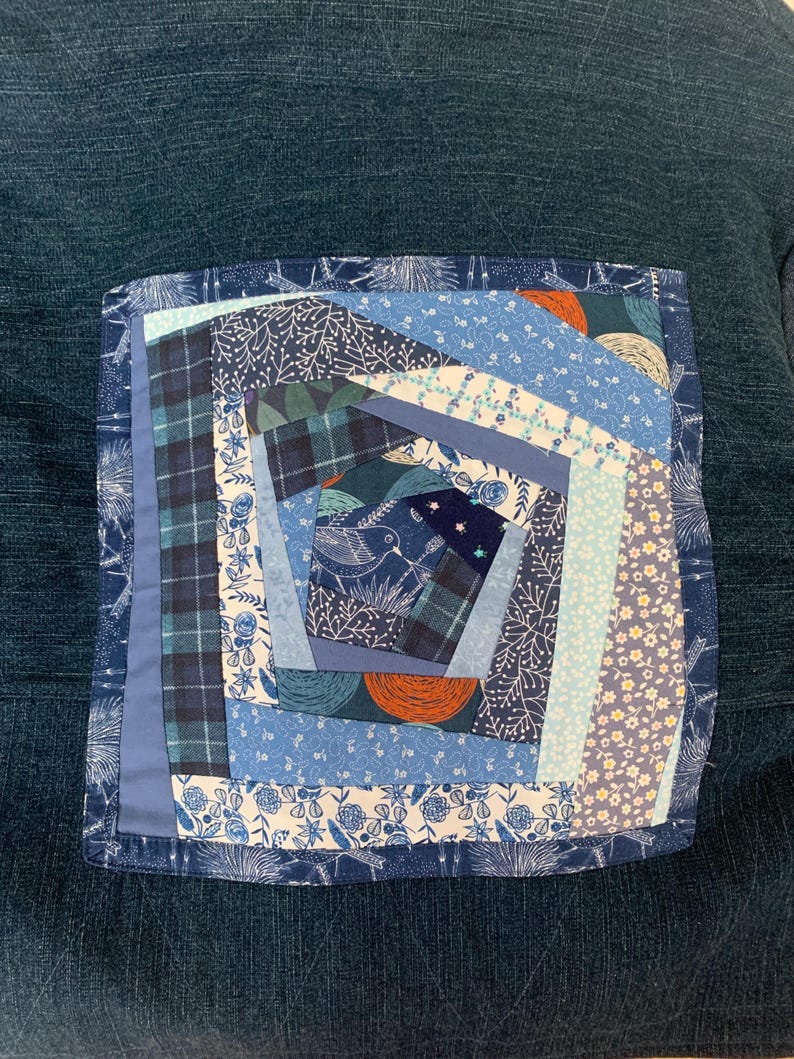 May include: A square fabric patch featuring a spiral patchwork design in various shades of blue, white, and orange. The quilt block includes patterns of flowers, plaid, and a bird illustration, all stitched onto a dark blue textured fabric base.