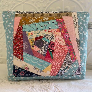 May include: A colourful patchwork fabric pouch with a spiral design. The pouch features floral and patterned fabrics in pink, blue, red, and purple. The edges are a light blue fabric with small white flowers.