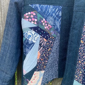 May include: A jacket constructed from dark blue denim fabric featuring a quilted front panel. The panel is composed of various fabric scraps in shades of blue, pink, and white, displaying floral, botanical, and bird patterns with visible stitching.
