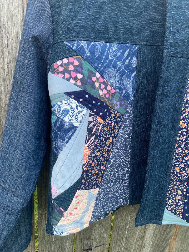 May include: A jacket constructed from dark blue denim fabric featuring a quilted front panel. The panel is composed of various fabric scraps in shades of blue, pink, and white, displaying floral, botanical, and bird patterns with visible stitching.