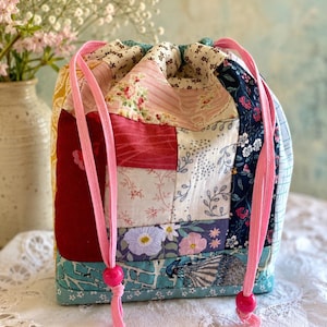 May include: A drawstring pouch constructed from various fabric patches featuring floral, grid, and solid patterns in shades of pink, red, navy blue, yellow, and teal. The bag includes two bright pink cords with matching pink beads at the ends.