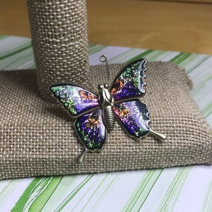 Puede incluir: Un broche de mariposa de plata con detalles de purpurina iridiscente en morado, verde y naranja.