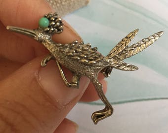 May include: A silver-toned brooch in the shape of a roadrunner with a turquoise stone on its head.