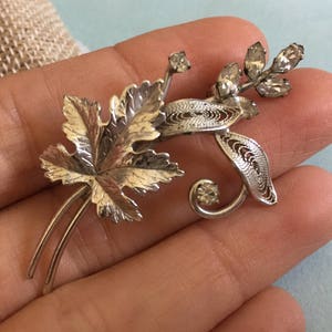May include: A silver filigree brooch with a leaf design and clear rhinestones.