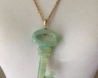 May include: A green jade key pendant necklace with a gold chain. The key is a large, ornate design with a detailed handle and a long, thin shaft.