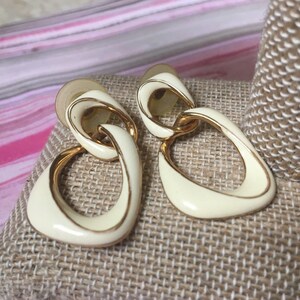 May include: A pair of white and gold dangle earrings with a geometric design. The earrings are made of metal and have a white enamel finish.