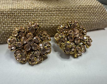 May include: A pair of gold-toned, flower-shaped clip-on earrings. Each earring features a cluster of light pink rhinestones set in a gold metal frame. The earrings are displayed on a neutral-colored surface.