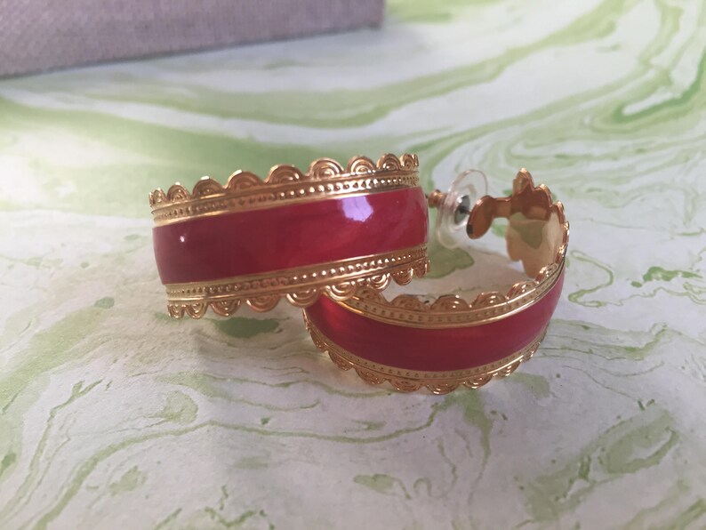 May include: A pair of gold hoop earrings with a red enamel design. The earrings have a scalloped edge and a delicate gold trim.