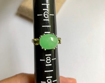 May include: A silver ring with a light green oval gemstone and two smaller green gemstones on the band. The ring is sized on a ring sizer that shows a size 8.