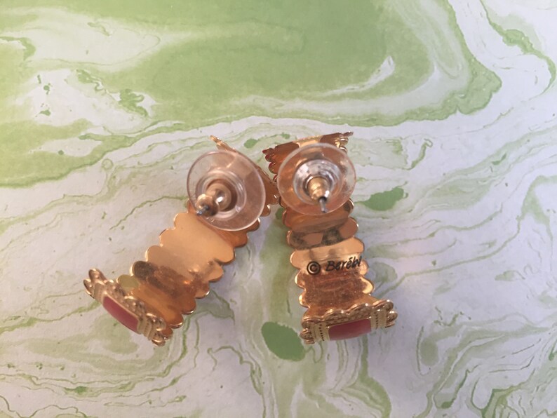 May include: A pair of gold hoop earrings with red accents. The earrings are marked with the word "Barozzi".