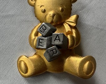 May include: A gold-colored teddy bear figurine holding gray letter blocks spelling "BEAR". The bear has a bow and is sitting with its legs extended. The figurine is made of metal.