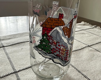 May include: A clear glass Coca-Cola tumbler featuring a winter village scene with snow-covered houses, a Christmas tree, and a sleigh. The glass has a classic, tall shape, perfect for cold beverages.