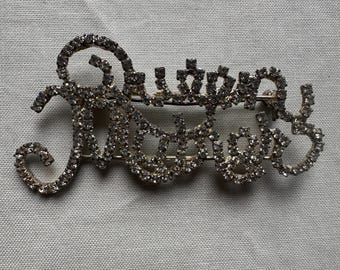May include: A silver-toned brooch spelling out "Queen" in a cursive font, encrusted with small, sparkling rhinestones. The brooch has a pin backing for secure attachment. The design is elegant and suitable for various occasions.