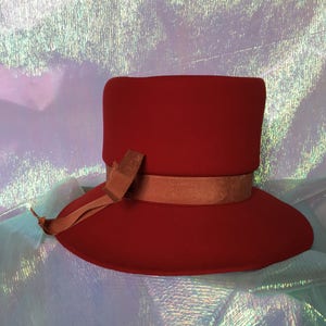 May include: A red felt hat with a wide brim and a brown ribbon. The hat is sitting on a iridescent background.