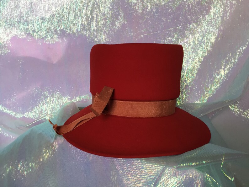 May include: A red felt hat with a wide brim and a brown ribbon. The hat is sitting on a iridescent background.