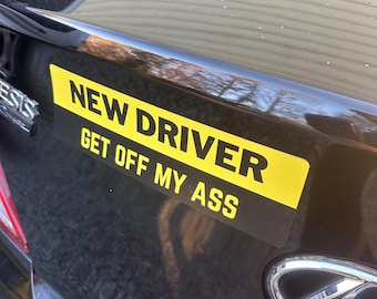 Könnte beinhalten: Ein schwarzes Auto mit einem gelb-schwarzen Aufkleber auf der Stoßstange mit der Aufschrift "NEW DRIVER GET OFF MY ASS". Der Aufkleber ist rechteckig und horizontal auf dem Kofferraum des Autos angebracht.