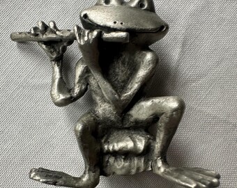 May include: A silver-colored figurine of a frog playing a flute. The frog is seated with its legs crossed and is holding the flute to its mouth with both hands. The figurine has a detailed, textured surface and is set against a gray background.