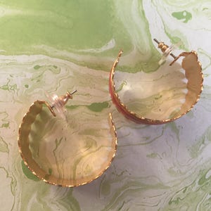 May include: A pair of gold hoop earrings with a scalloped edge and a red enamel stripe. The earrings are on a white background with green marbling.