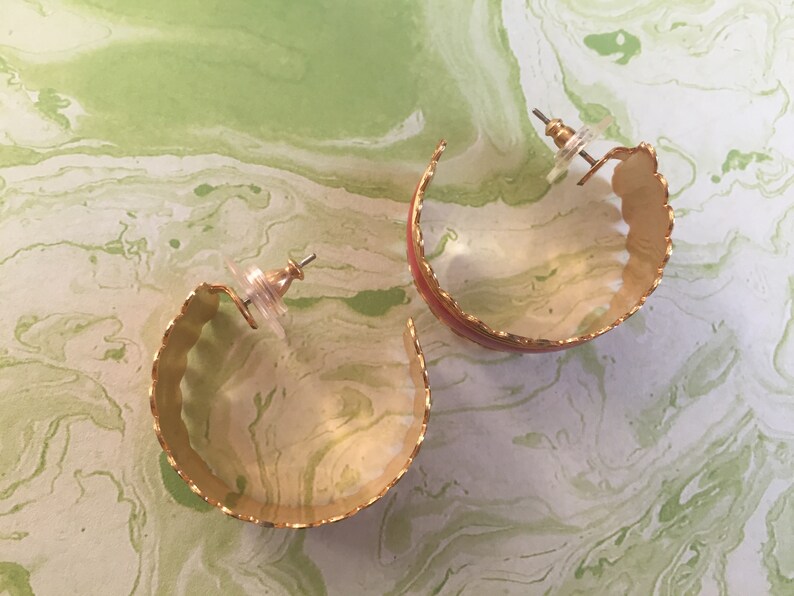 May include: A pair of gold hoop earrings with a scalloped edge and a red enamel stripe. The earrings are on a white background with green marbling.