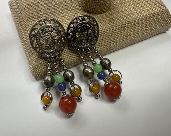 May include: Silver-toned filigree earrings with dangling beaded accents. Each earring features a circular design with intricate patterns, and colorful beads in green, blue, yellow, and red. The earrings are displayed on a neutral-toned surface.