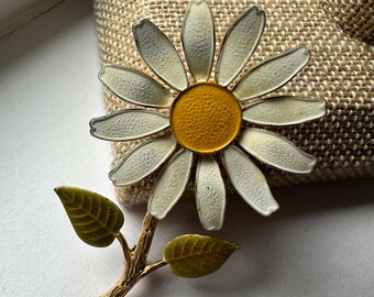 May include: A vintage enamel daisy brooch with a yellow center and white petals. The brooch has a gold stem with two green leaves.