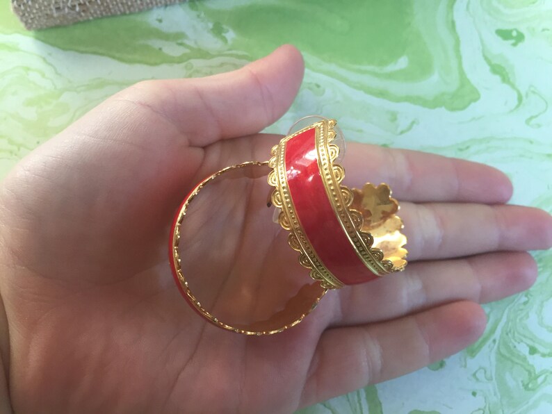 May include: A pair of gold hoop earrings with a red enamel band. The earrings have a decorative scalloped edge.