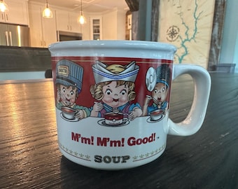 May include: White ceramic mug with a red rim and handle. The mug features three cartoon characters enjoying soup, with the text "M'm! M'm! Good!" and "SOUP" printed on the front. The characters wear hats.