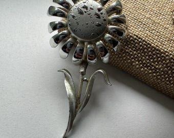 May include: A silver-toned brooch in the shape of a flower with a round center and a stem with leaves.