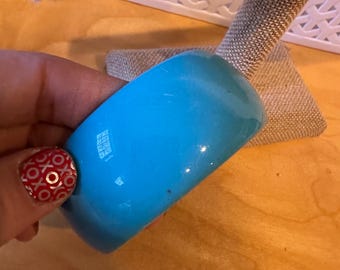 May include: A bright turquoise bangle bracelet. The smooth, rounded bracelet is a solid color and appears to be made of a glossy material. The bracelet is held in a hand, and the background is a light brown surface.