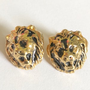 May include: A pair of gold lion head earrings with black enamel details. The lion heads are facing forward with their mouths open.