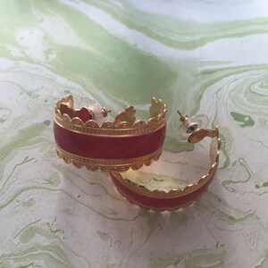 May include: A pair of gold hoop earrings with a red enamel band and a decorative gold trim. The earrings are on a white and green marbled surface.