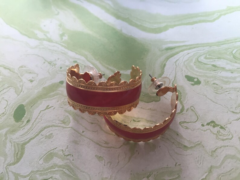May include: A pair of gold hoop earrings with a red enamel band and a decorative gold trim. The earrings are on a white and green marbled surface.