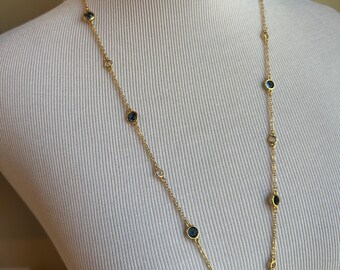 May include: A long gold-colored necklace with alternating dark blue and clear circular stones. The necklace is draped on a white mannequin, showcasing its length and design. The chain is delicate, and the stones are evenly spaced.