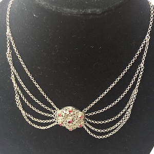 Op de afbeelding: Een zilveren ketting met vier kettingen en een rood strass-middelpunt.