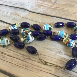 May include: A blue and white ceramic bead necklace with a brass chain. The beads are oval shaped and have a painted design of houses and trees.
