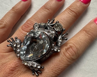 May include: A large, silver-toned frog ring with a large, clear, teardrop-shaped crystal as the frog's body. The ring is adorned with smaller clear crystals and black accents. The frog's legs and eyes are detailed. The background is a light fabric.
