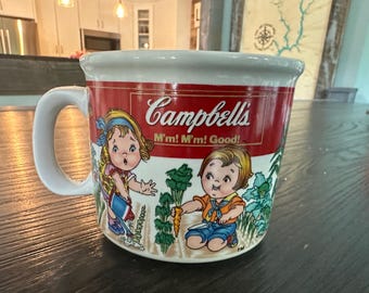 May include: A white ceramic Campbell's soup mug with a red band featuring the Campbell's logo and the phrase "M'm! M'm! Good!". The mug has illustrations of children in a garden.