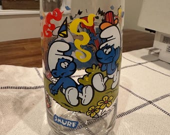 May include: A clear glass featuring a colorful design of Smurfs. The glass has a cylindrical shape with a Smurf logo at the bottom. The design includes blue Smurf characters, yellow and red decorative elements, and green grass.